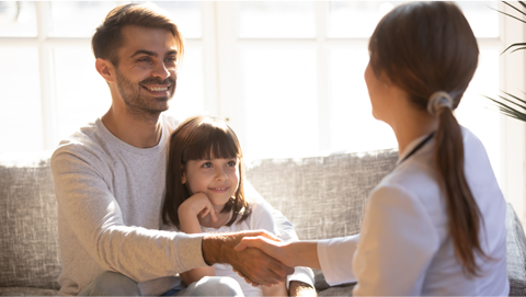 Female child sitting on adult male's lap on a couch. Adult female in foreground shaking man's hand.