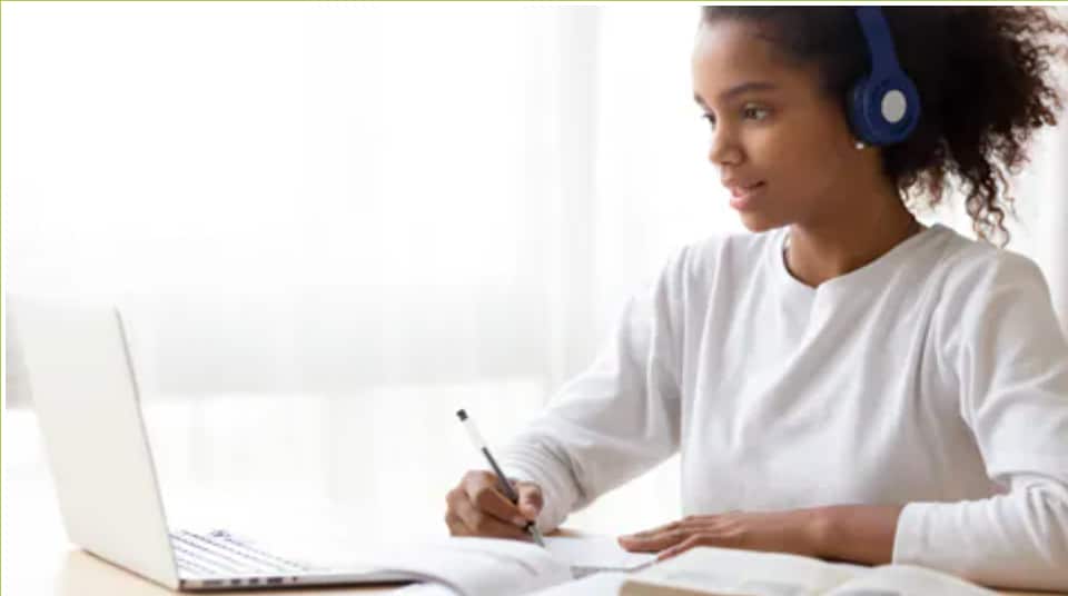 Adolescent female wearing headphones and writing on paper while sitting at a desk and looking at a laptop computer.