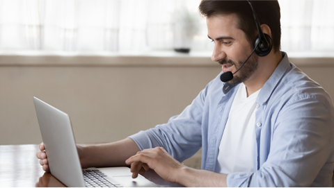 Adult man wearing headphones with microphone sitting at desk using a laptop computer.
