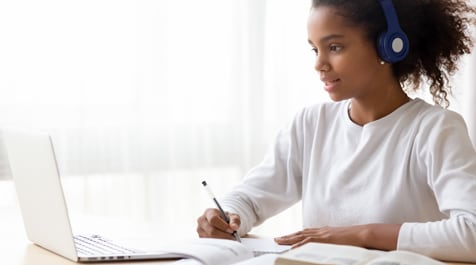Adolescente portant un casque d’écoute et écrivant sur une feuille de papier; elle est assise à un bureau devant un ordinateur portable.